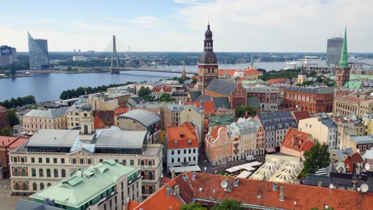 Panorāmas skats uz Rīgas vecpilsētu ar Daugavas upi un tiltiem saulainā dienā.