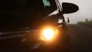 Tumšs automobilis ar ieslēgtiem dzelteniem lukturiem miglainā rudens rītā uz ceļa.