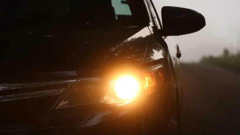 Tumšs automobilis ar ieslēgtiem dzelteniem lukturiem miglainā rudens rītā uz ceļa.