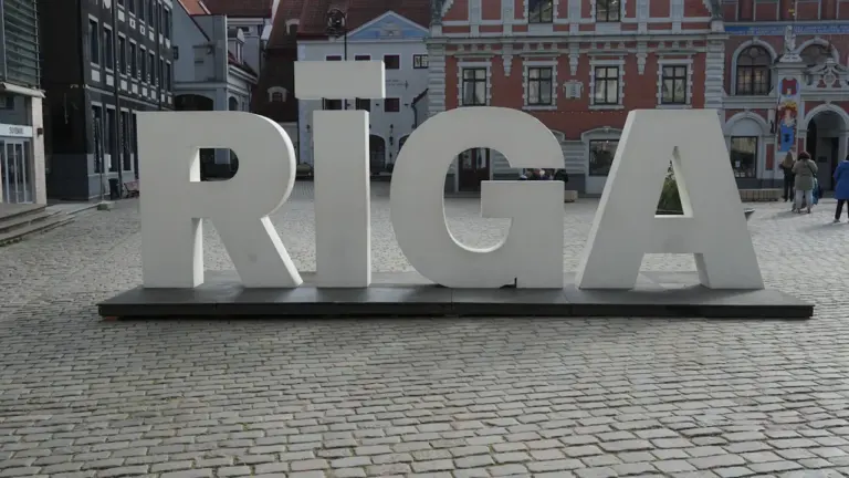 Liels uzraksts RIGA Rīgas vecpilsētas bruģētā laukumā ar vēsturiskām ēkām fonā.