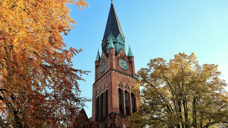 Baznīcas tornis rudens noskaņās ar zeltainām koku lapām skaidrā zilā debesī.