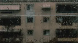 Lietus pilieni uz dzīvokļa loga stikla rudenīgā laikā, fona redzama ēka.