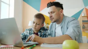 Tēvs palīdz dēlam pildīt mājasdarbus pie datora mājās, smaidot un mācoties kopā.