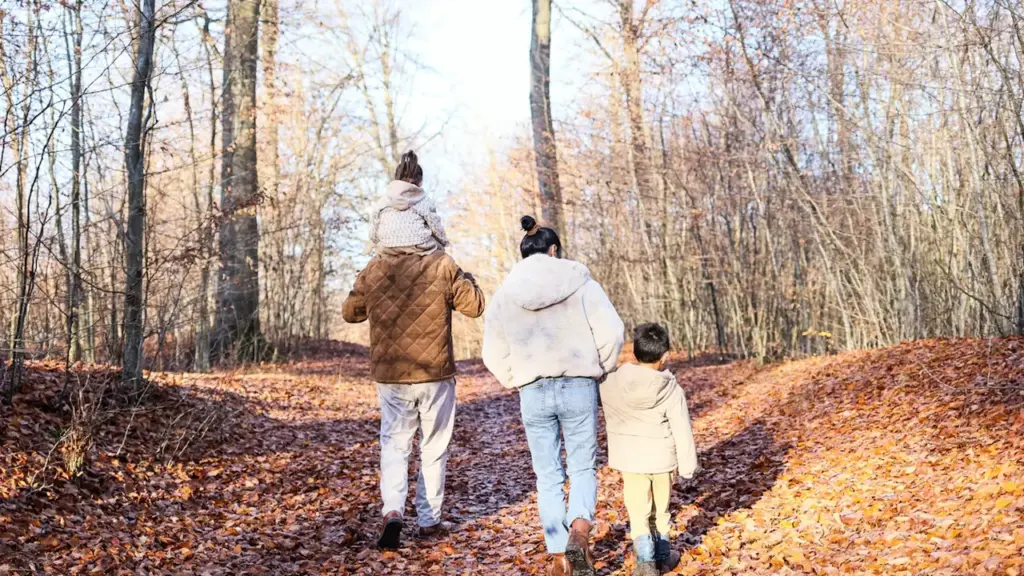 Ģimene ar bērniem pastaigājas pa rudens meža taku klātu ar lapām.